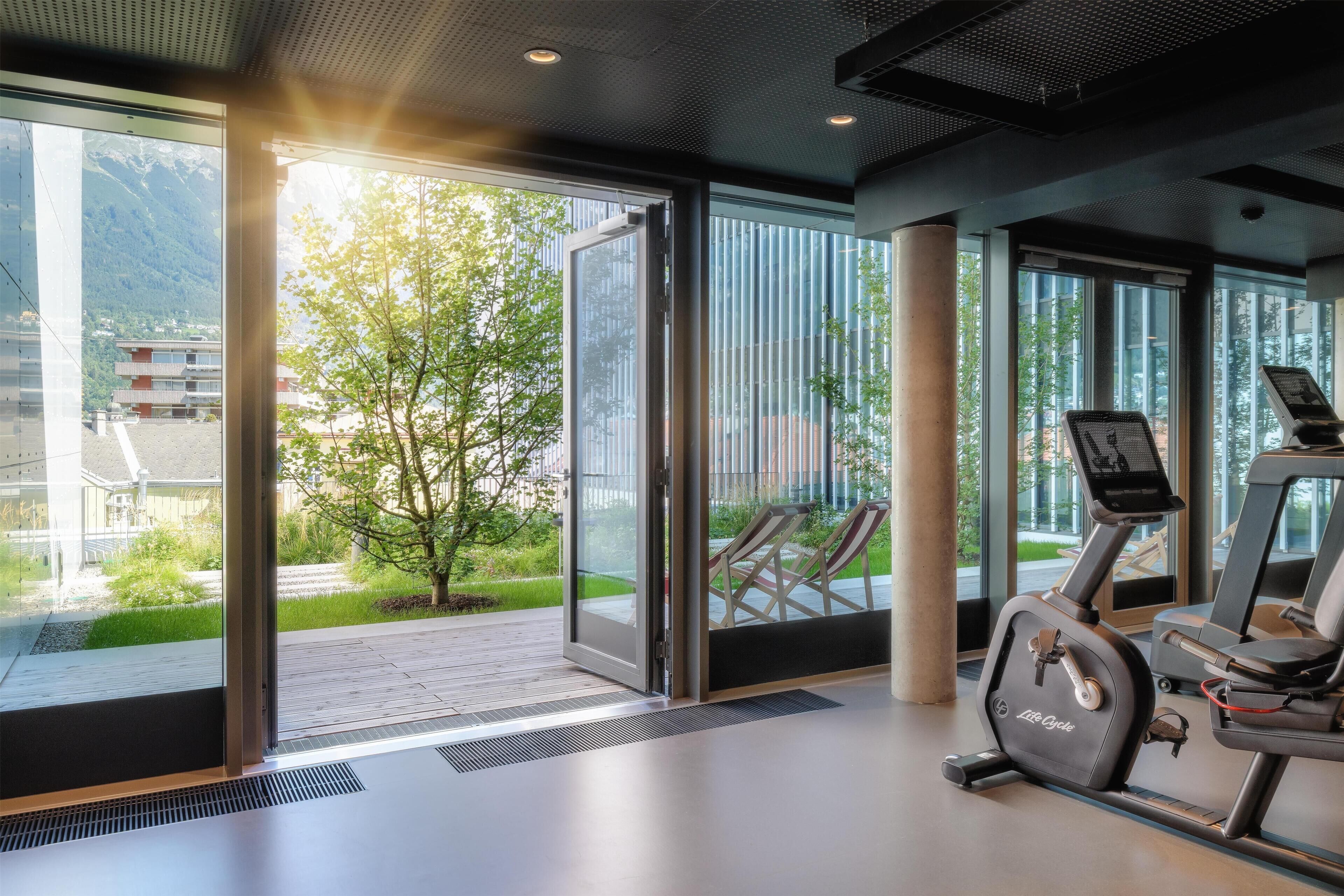Une salle de sport équipée de matériel d’exercice et dotée de grandes fenêtres offrant une vue sur la verdure et les bâtiments.