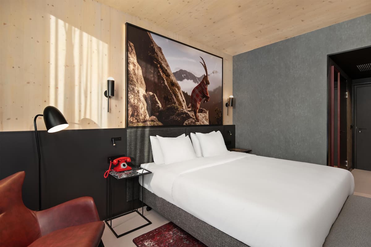 Ein Hotelzimmer mit einem großen Bett, einem roten Telefon, einer Schreibtischlampe, einem roten Sessel und einem Wandbild mit einer Berglandschaft.