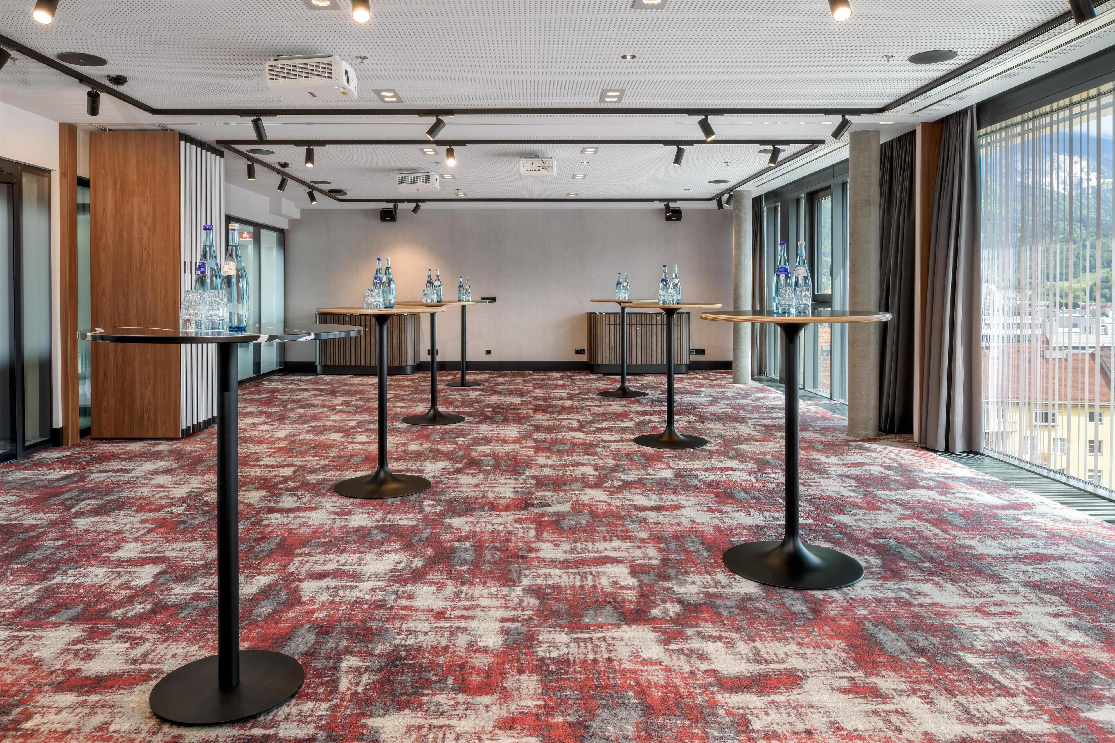 Un salón de conferencias con alfombra roja estampada, varias mesas con taburetes y ventanas amplias que ofrecen vistas.