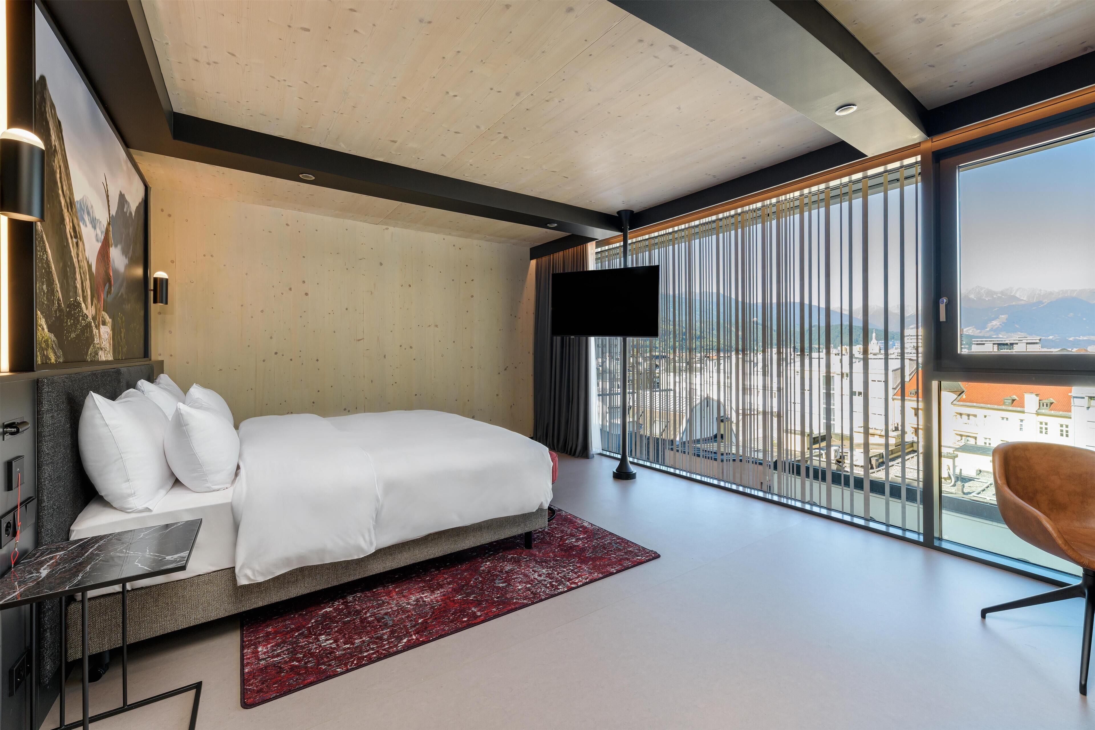 A modern hotel room with a large bed, a flat-screen TV, a view of the city, and a balcony with a chair.