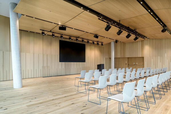 A conference room with rows of white chairs, a large screen, and wooden panel walls.