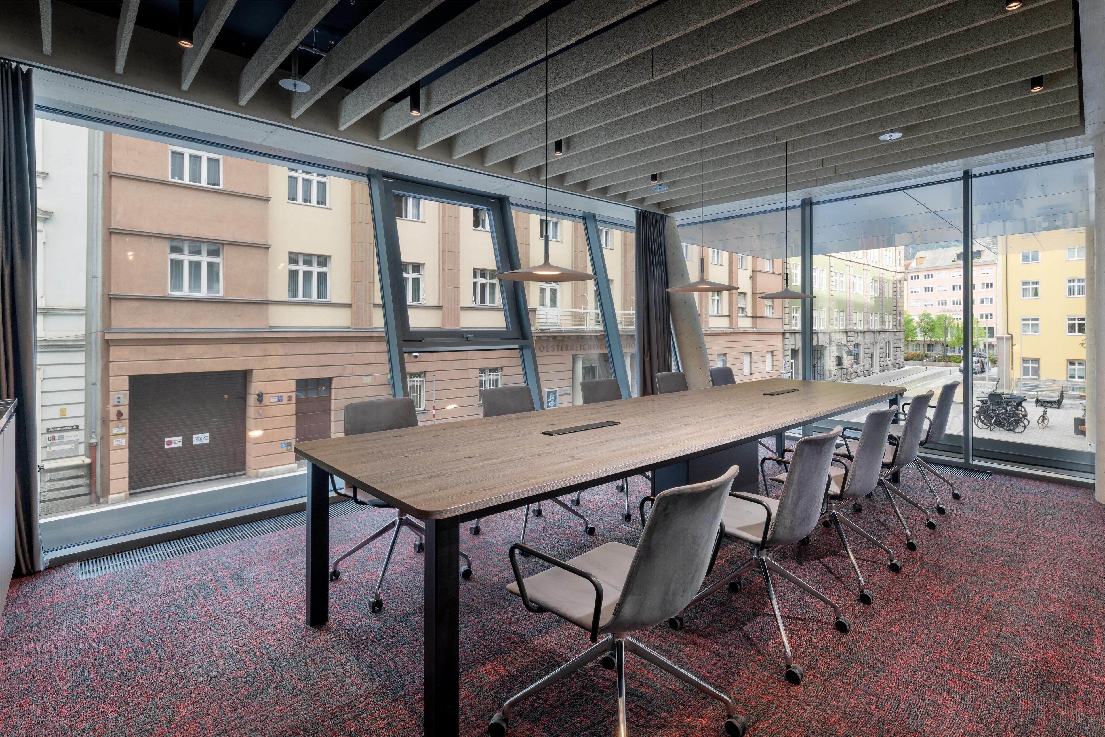 A conference room with a long table, chairs, and large windows offering a view of buildings outside.