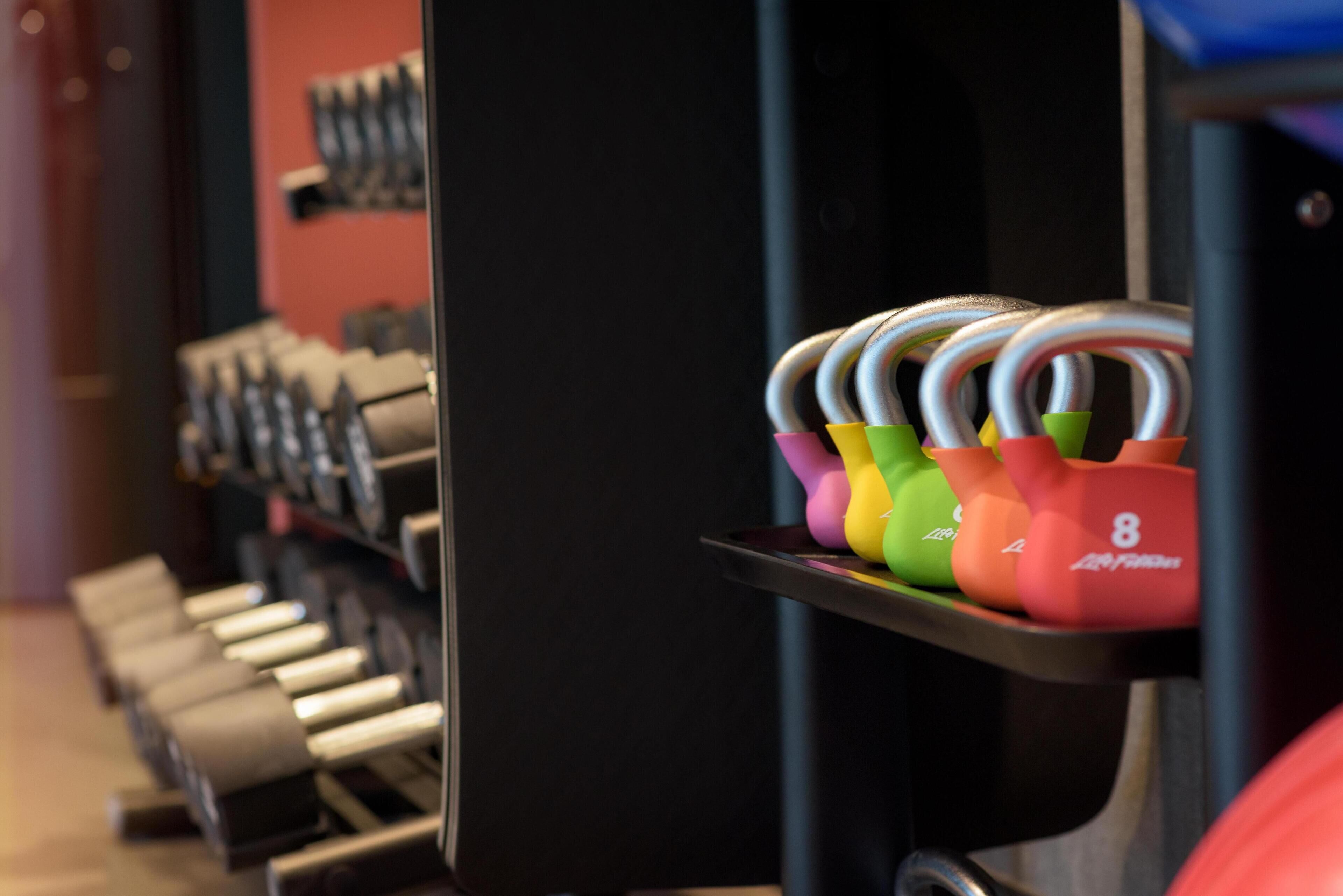 Eine Reihe von Kurzhanteln auf einem Gestell, ein schwarzes Schranks und ein Regal mit farbenfrohen Kettlebells.