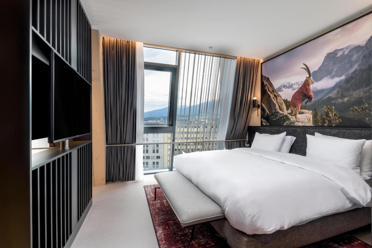 Ein modernes Hotelzimmer mit einem großen Bett, einem Flachbildfernseher, einer Sitzbank und Blick auf die Stadt.