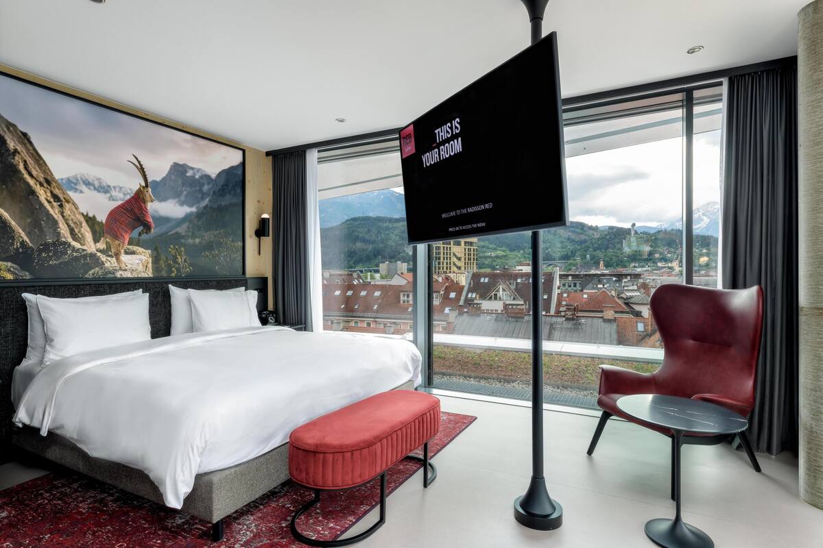 Ein modernes Hotelzimmer mit einem großen Bett, einem roten Sessel, einem kleinen Tisch und einem Fernseher, der eine Willkommensbotschaft zeigt.