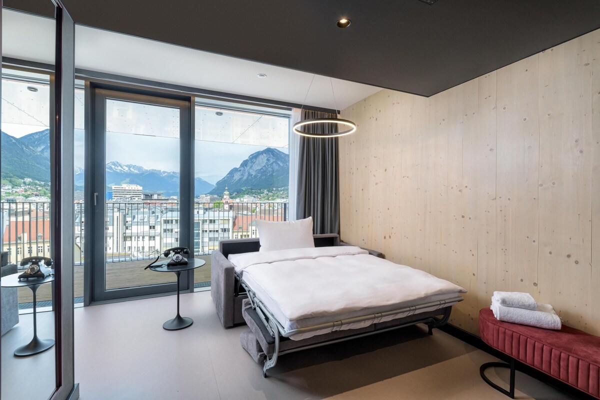 A modern hotel room with a bed, a small table with a telephone, a bench with towels, and a large window offering a mountain view.