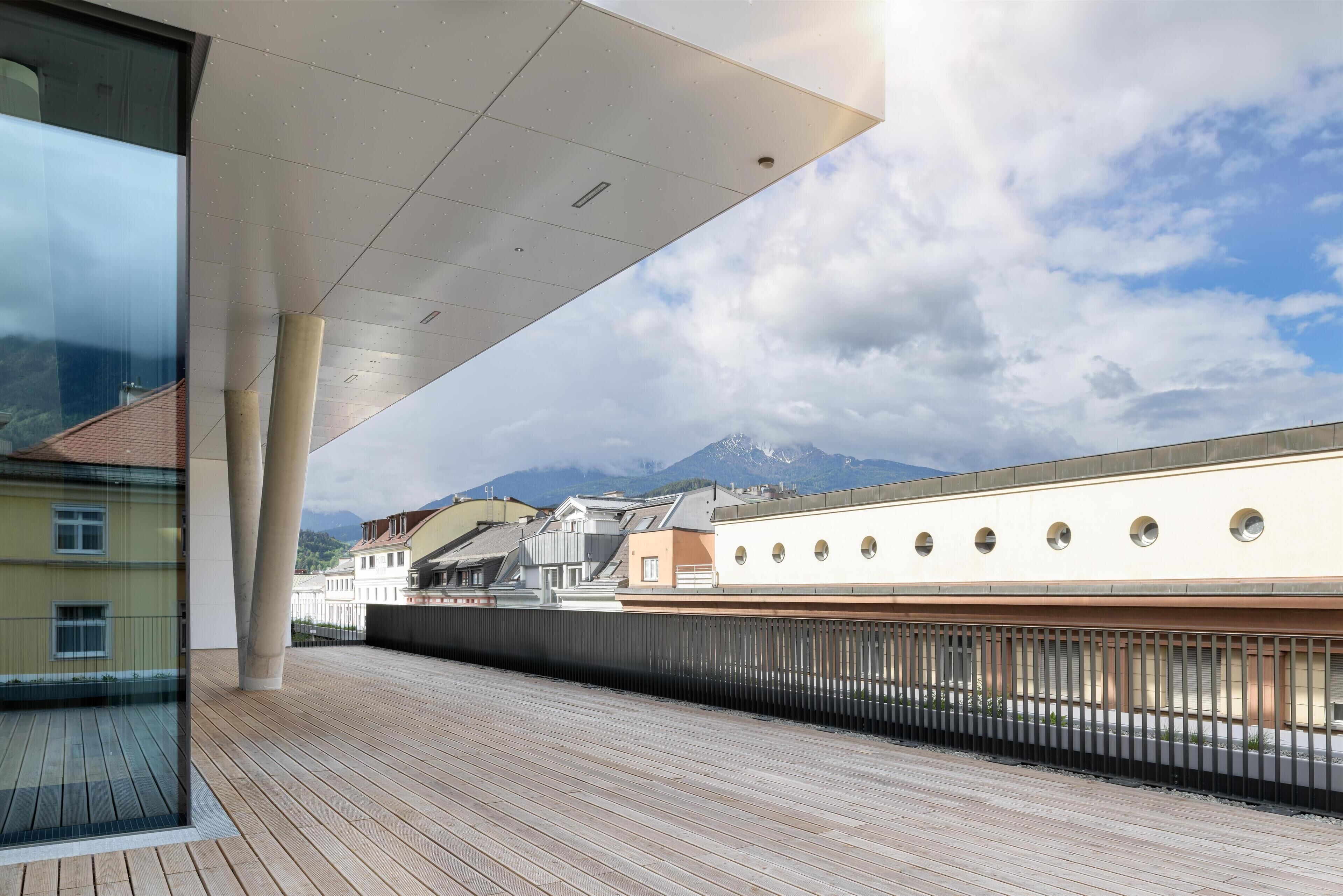 A modern building with a wooden deck, large windows, and a view of mountains.