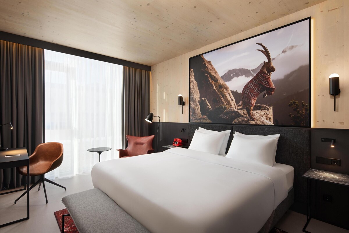 A hotel room with a large bed, a desk, a chair, a nightstand, and a wall-mounted TV.