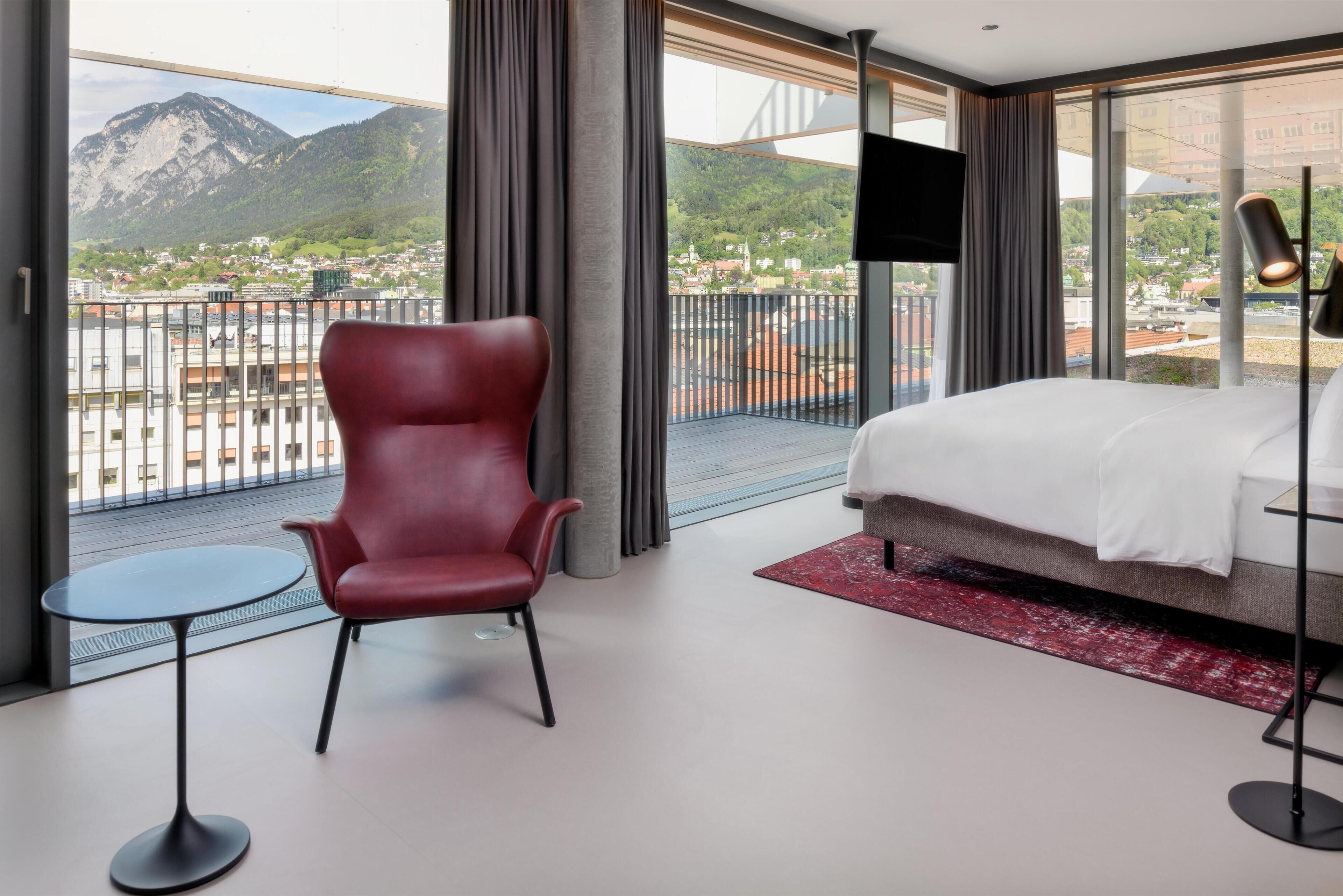 Une chambre d’hôtel avec un lit, une chaise rouge, une petite table, un téléviseur et un balcon offrant une vue sur la montagne.