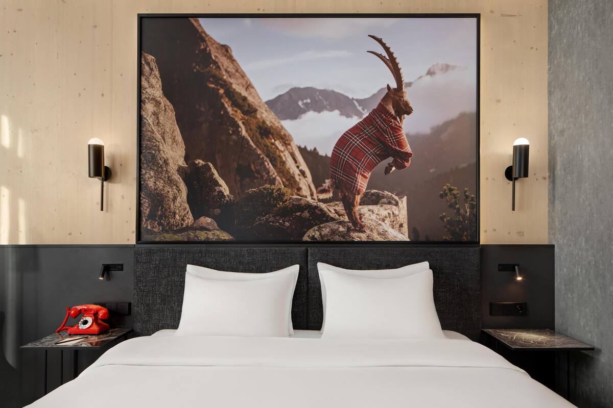 Ein Hotelzimmer mit einem Bett, einem Wandbild von einer Berglandschaft, einem roten Wählscheibentelefon und zwei Wandleuchten.