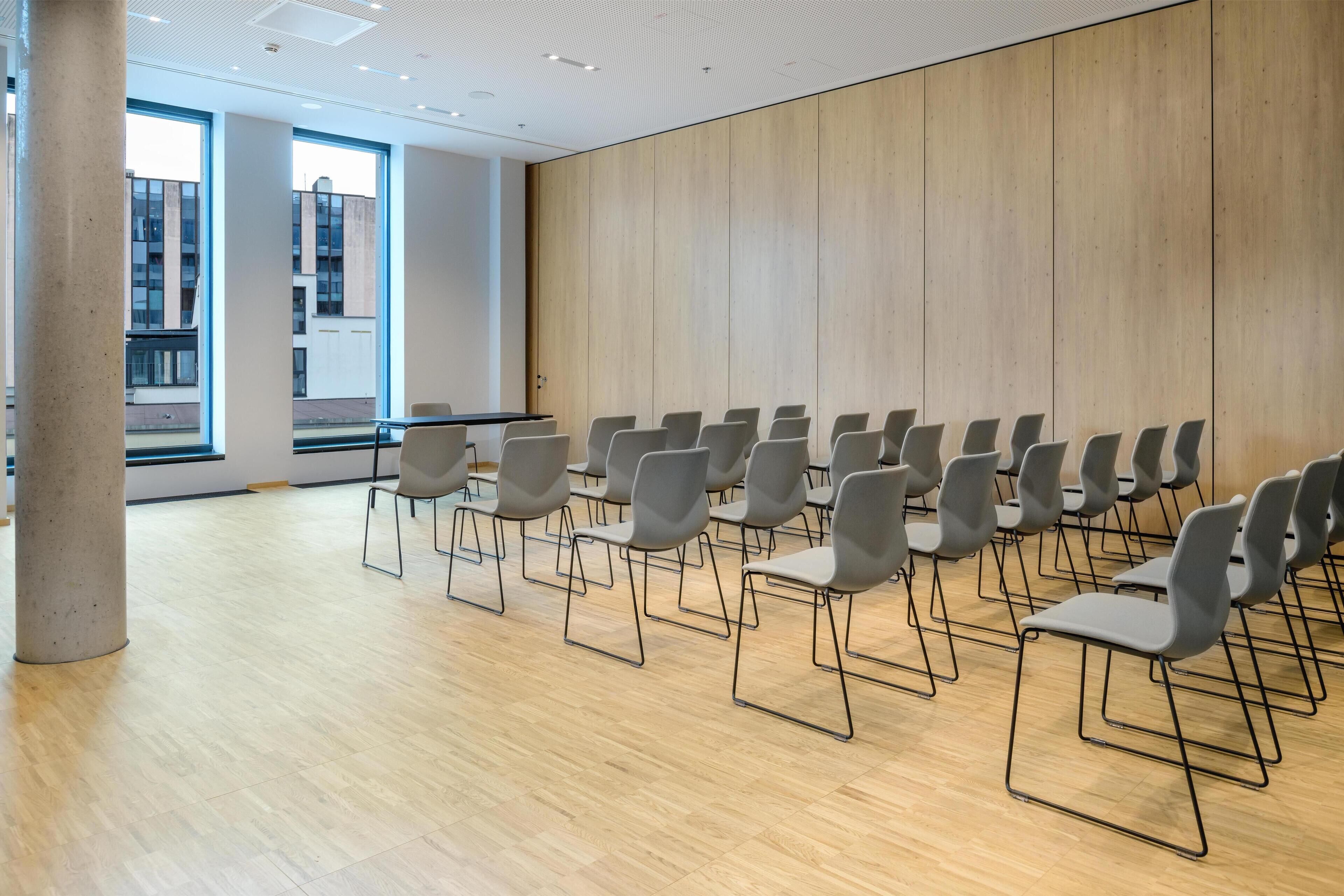 Une salle de conférence avec des rangées de chaises, de grandes fenêtres et un mur en bois.