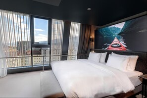 A hotel room with a large bed, a view of the city through floor-to-ceiling windows, and a painting of a kayak on the wall.
