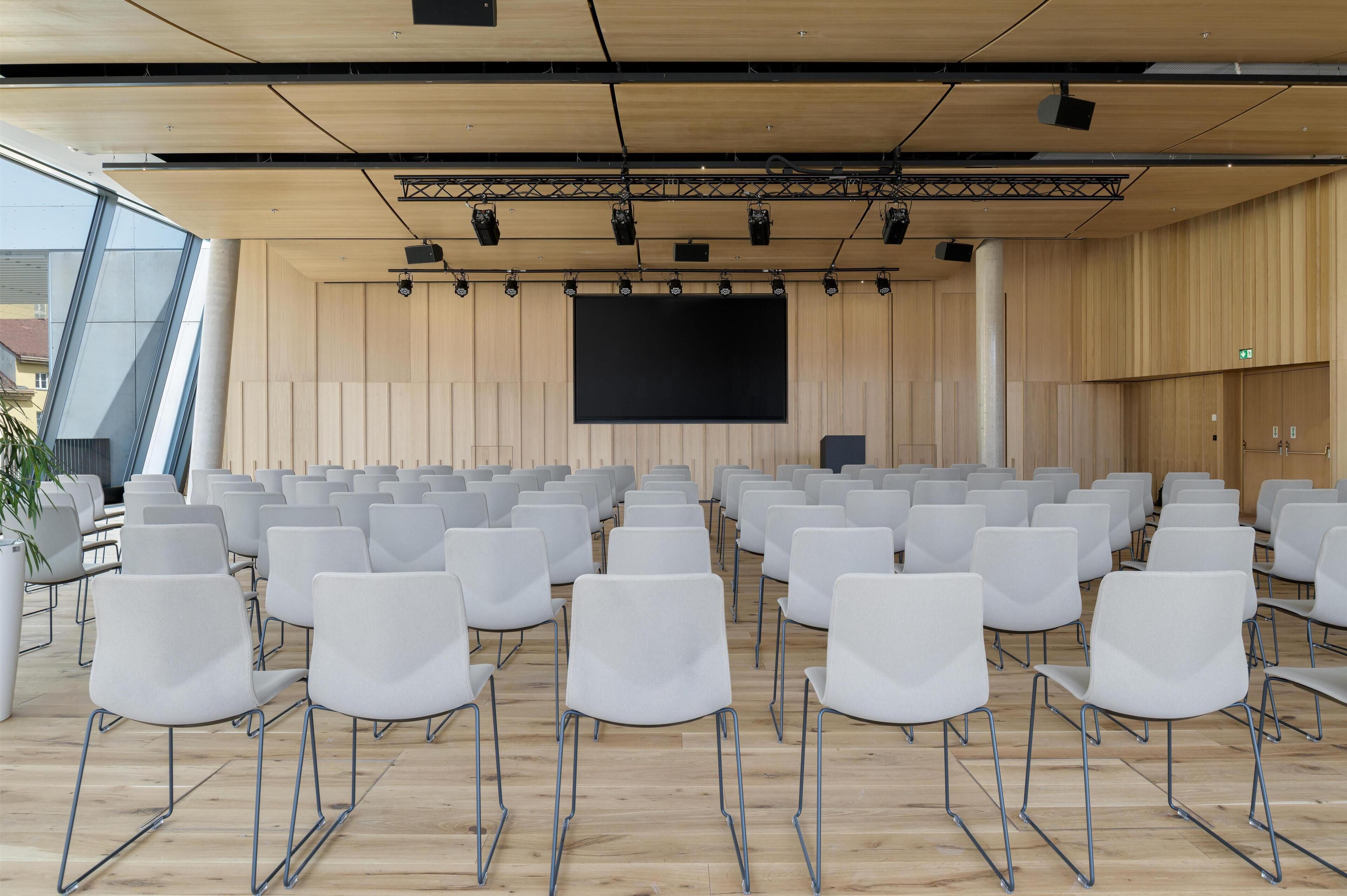 A conference room with rows of white chairs, a large screen, and wooden paneling.