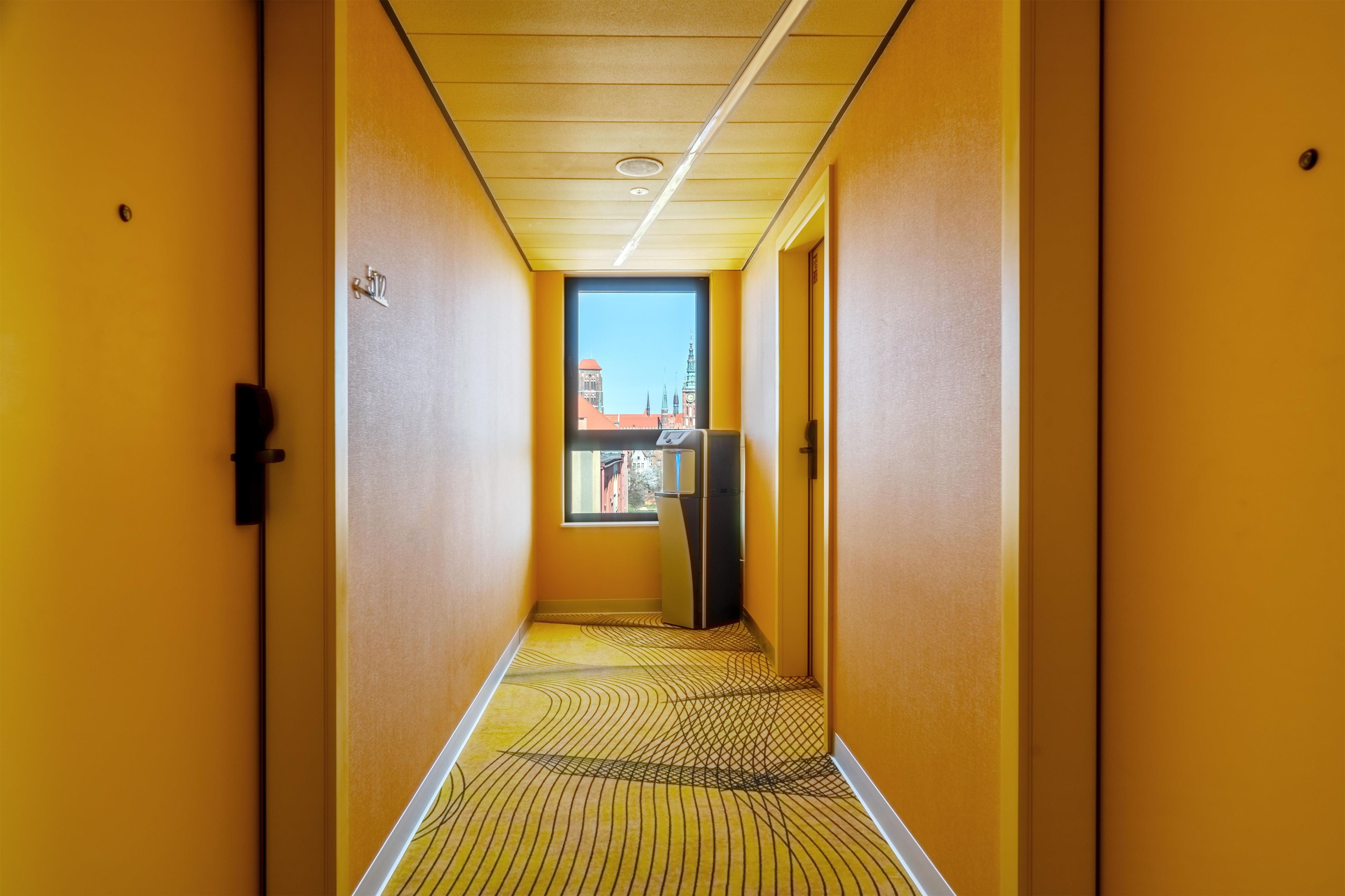 Un couloir étroit aux murs jaunes et au sol à motifs, qui mène à une porte donnant sur des bâtiments à l’extérieur.