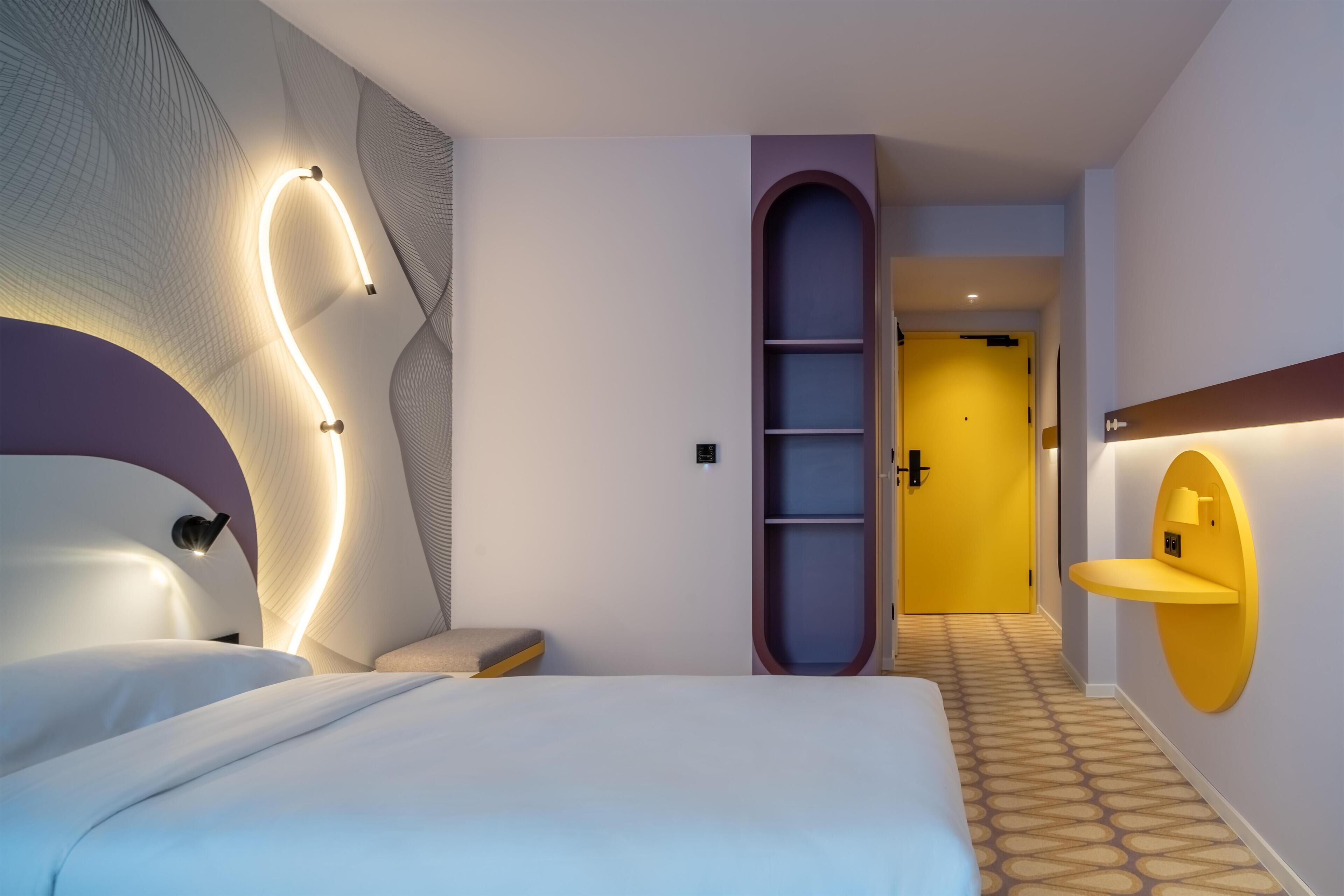 Une chambre d’hôtel moderne dotée d’un grand lit, d’un luminaire incurvé encastré, d’un petit coin salon et d’un couloir menant à une porte.