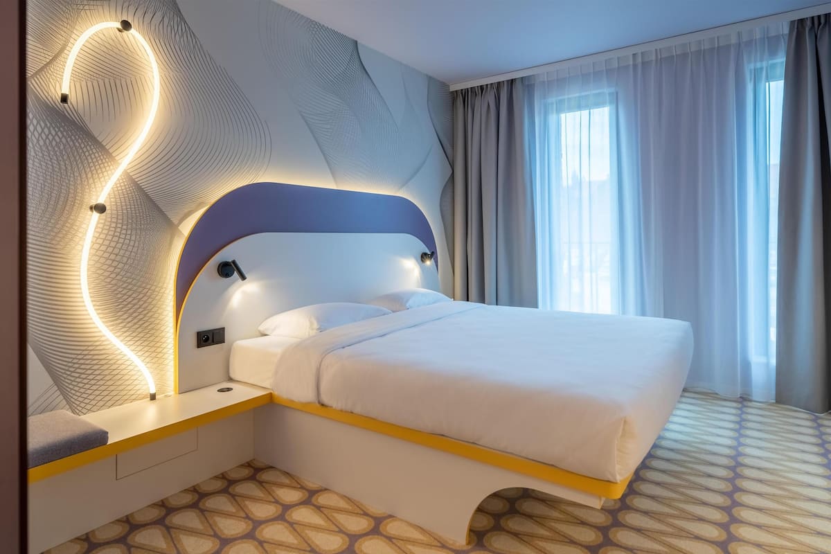 A modern hotel room with a curved headboard, a bed with white linens, a patterned carpet, and a large window with curtains.