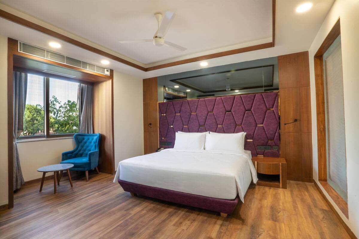 A modern hotel room with a large bed, a blue armchair, a small table, and a ceiling fan.
