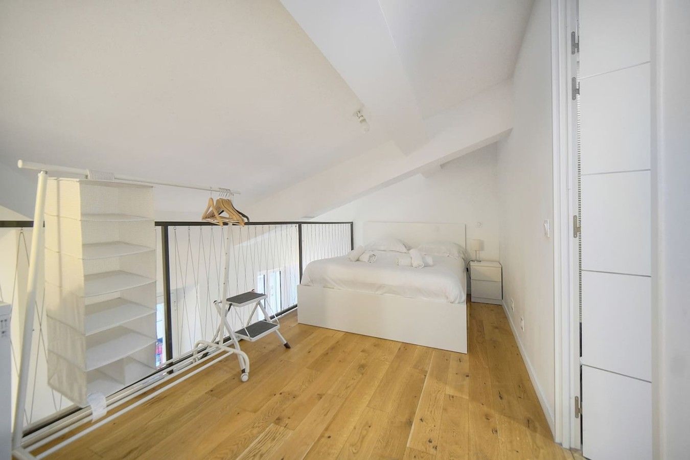 A bedroom with a bed, a nightstand, a white wardrobe, and a wooden floor.