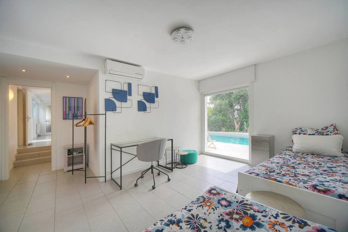 A modern bedroom with a desk, two beds, a view of a swimming pool, and a door leading to a bathroom.