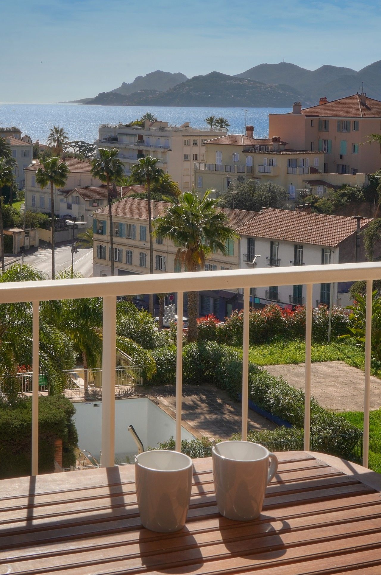 A balcony with two cups, overlooking a pool and a view of buildings and mountains.