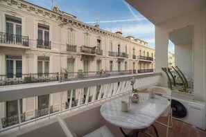 A balcony with a white railing, a table with a fan, and a view of pastel-colored buildings. - 13232804 FRGK Victoria 3 Central Cannes Studio Ter (Cannes)