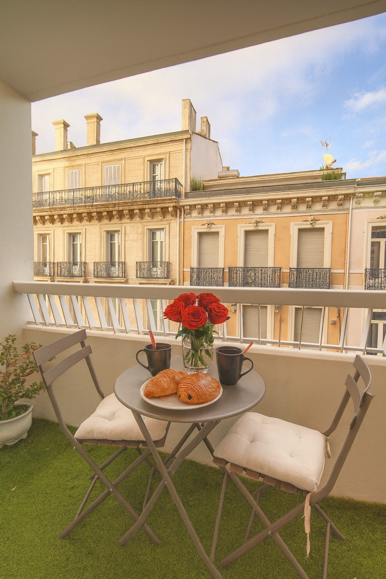 Un balcone con un tavolo apparecchiato per due persone, con un vaso di rose rosse e croissant.