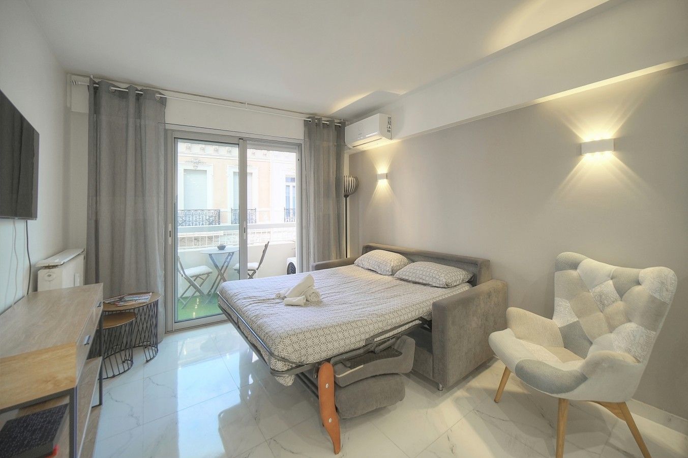 A modern hotel room with a bed, a chair, a desk, and a balcony with a table and chairs.