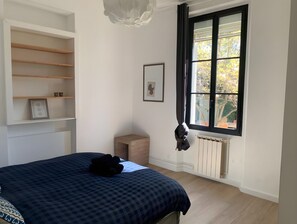 Une chambre à coucher avec un lit, une fenêtre avec des rideaux, un radiateur et un petit tabouret en bois.