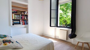 Ein Schlafzimmer mit einem Bett, einem Bücherregal, einem Heizkörper, einem Fenster mit Blick auf Bäume und einem weißen Stuhl.
