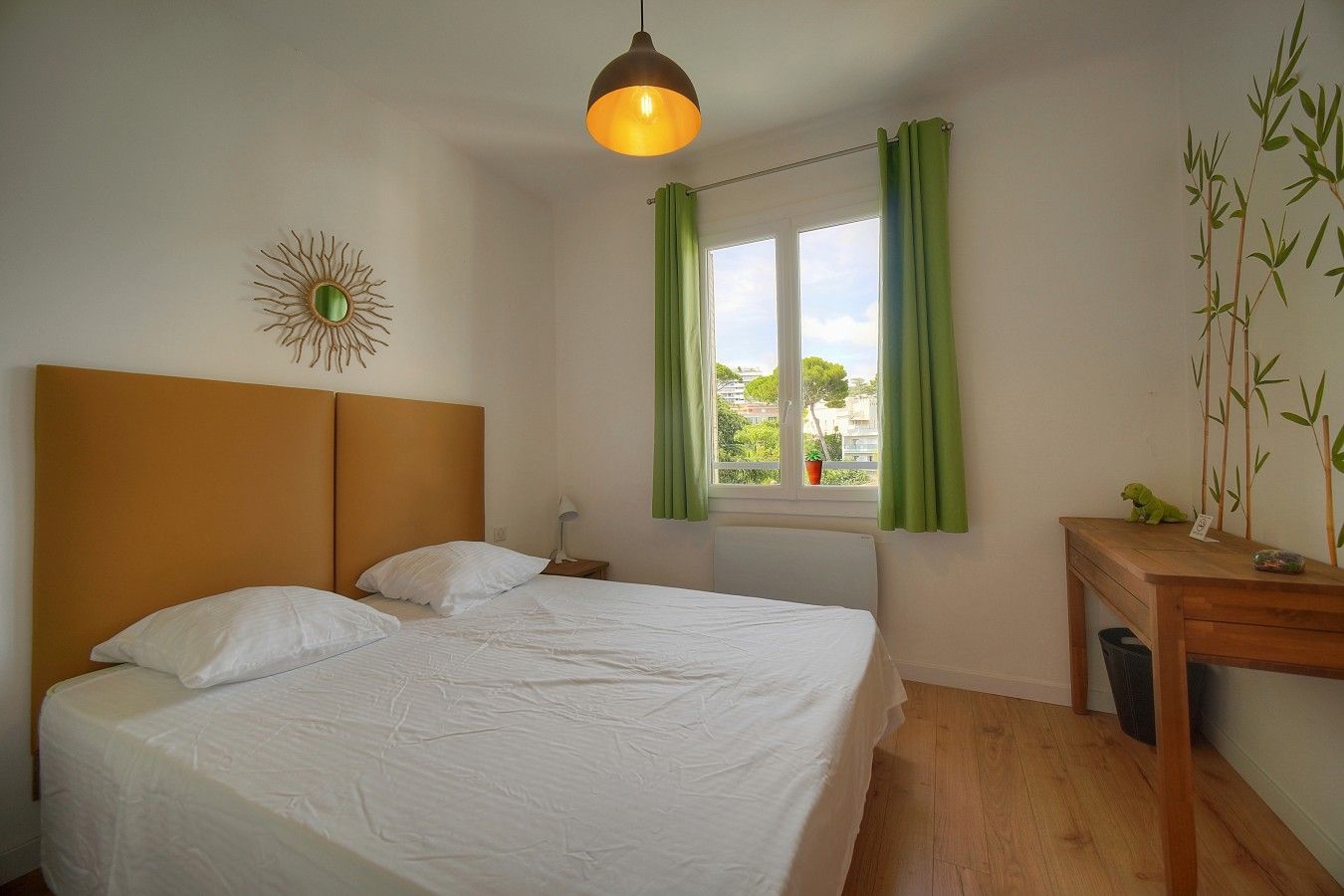 Quarto com cama, mesa de cabeceira, abajur, janela com cortinas e um objeto decorativo na parede.