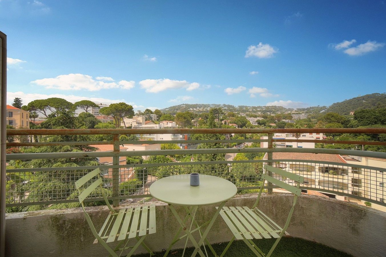 Un balcon avec une table et des chaises, donnant sur un quartier résidentiel bordé d’arbres et de bâtiments.