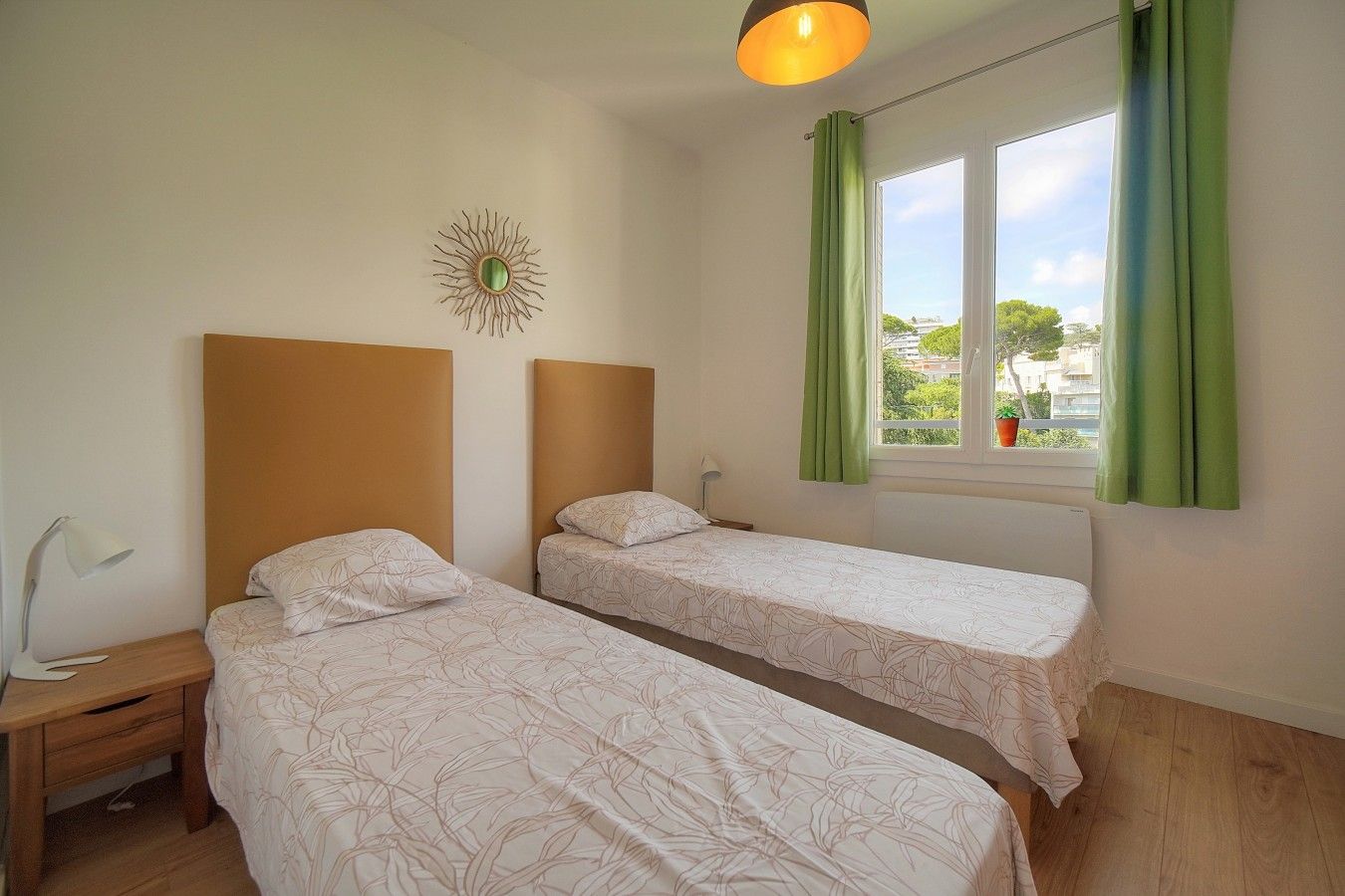 A room with two single beds, a bedside table with a lamp, a window with curtains, and a wall decoration.