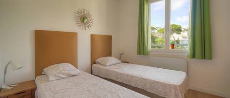 A room with two single beds, a bedside table with a lamp, a window with curtains, and a wall decoration.