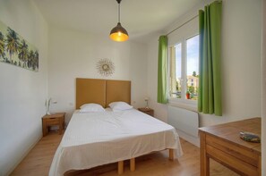 A bedroom with a bed, bedside tables, a desk, a hanging light fixture, a window with curtains, and a wall decoration. - 12251546 FRGK Isola Bella ` 2-Bedroom, High-Speed Wi-Fi AC ` (Cannes)