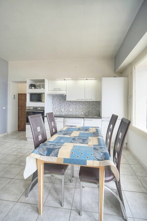 Comedor con una mesa dispuesta para seis personas, rodeada de sillas, en una habitación con una cocina al fondo.
