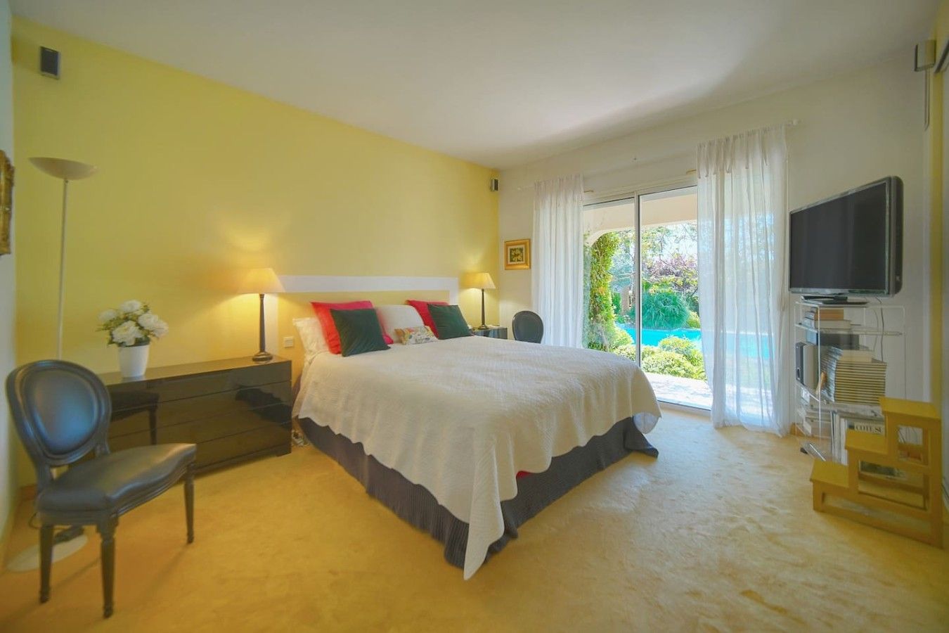 Un dormitorio con cama, mesitas de noche, una silla, televisor y vistas a un jardín.