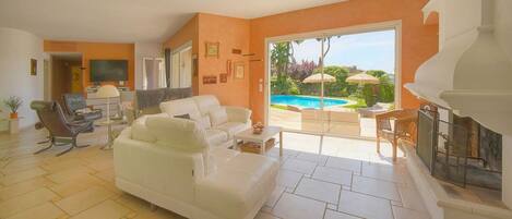 A spacious living room with a white sofa, a television, and a fireplace. There is a large sliding glass door leading to an outdoor area with a pool and umbrellas.