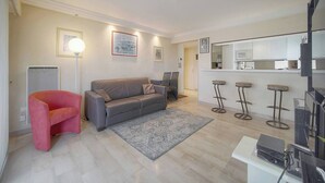 A modern living room with a grey sofa, a red armchair, a grey rug, and a kitchen area with a microwave and bar stools. - ATHENA (Cannes)