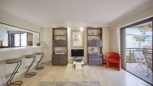 A modern living room with a TV, bar stools, a red chair, and a balcony with a view. - ATHENA (Cannes)