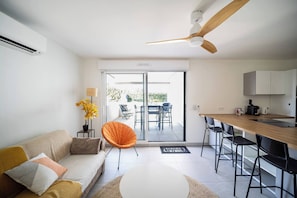 A modern living room with a sofa, a dining table, a kitchen area, and a sliding glass door leading to an outdoor patio. - BEL AZUR (Antibes)