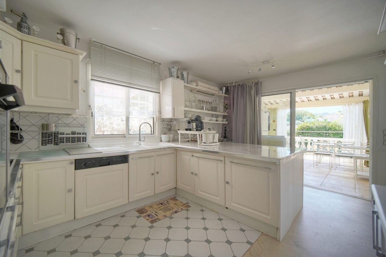 Una cocina moderna con muebles blancos, lavavajillas y un ventanal con persianas.