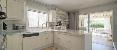 Una cocina moderna con muebles blancos, lavavajillas y un ventanal con persianas.