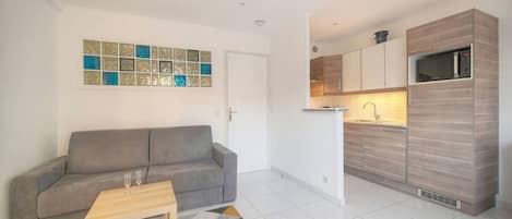 A compact living space with a sofa, a small table, a kitchen with modern appliances, and a decorative wall.
