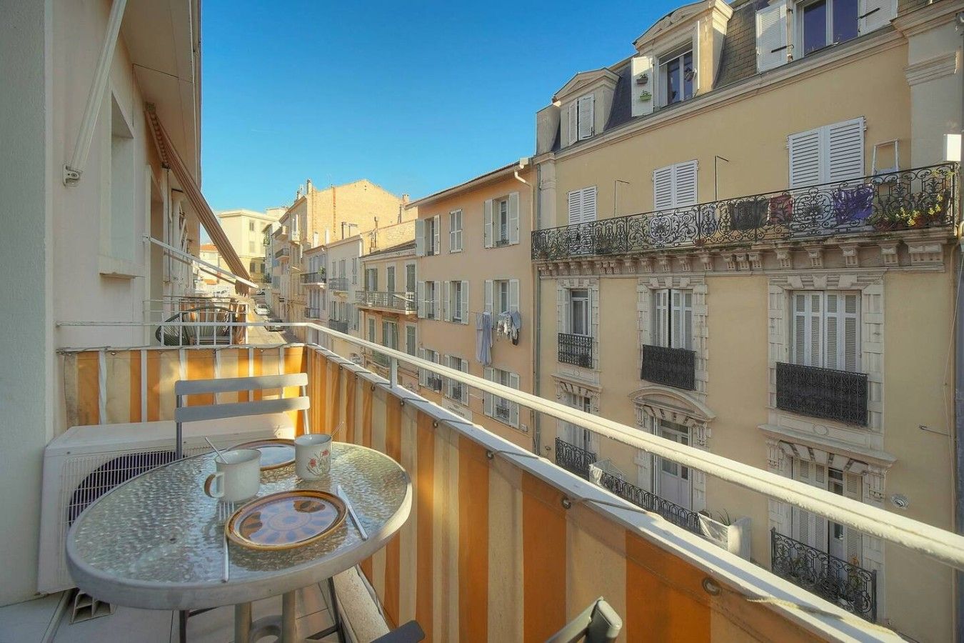 Un balcon avec une table et des chaises, donnant sur une rangée d’immeubles dotés de balcons et de volets.