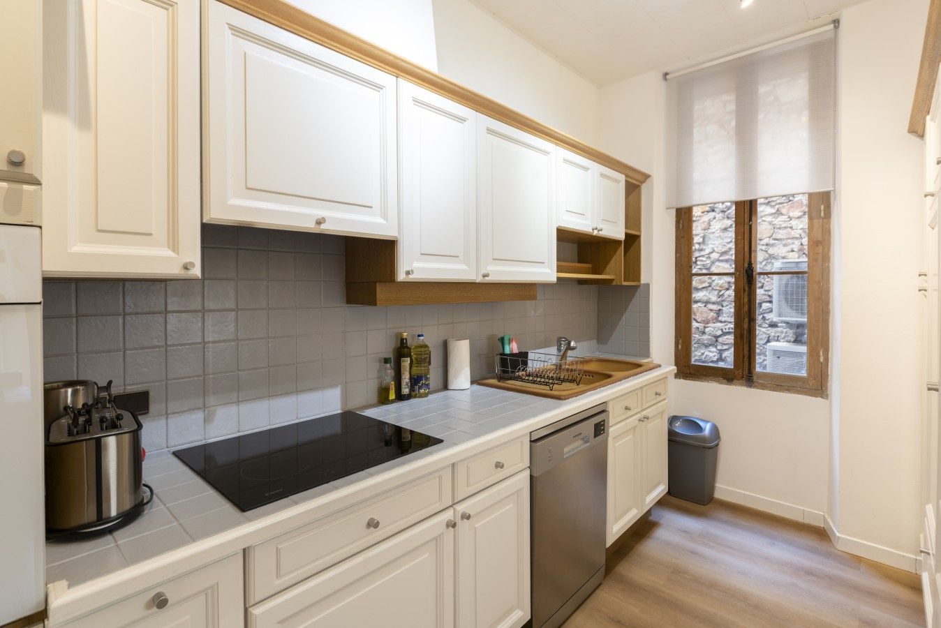Cocina con armarios blancos, placa de inducción negra, lavavajillas y ventana con vista a una pared de piedra.