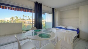 Une chambre d’hôtel avec un balcon, un lit, une table ronde avec des chaises, et une vue sur les palmiers et les immeubles.