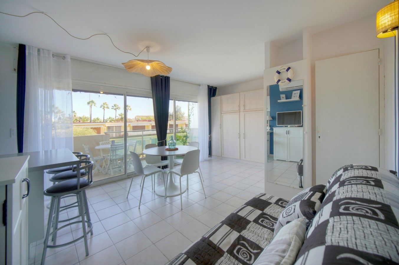 A modern hotel room with a balcony, a dining area, a kitchenette, and a bed with patterned bedding.