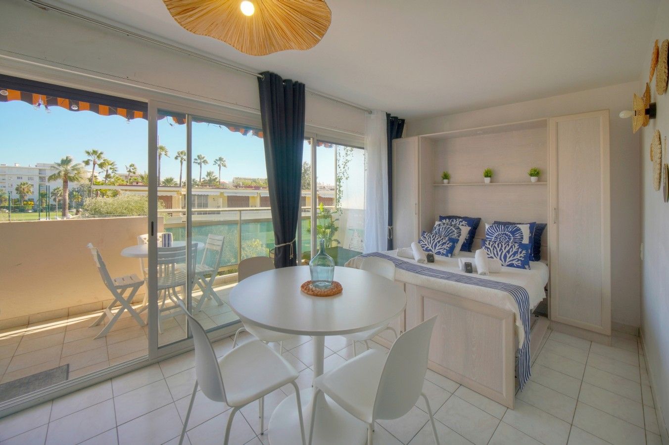 A balcony with a table and chairs, a bed with blue and white bedding, and a view of palm trees.
