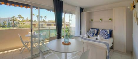 A balcony with a table and chairs, a bed with blue and white bedding, and a view of palm trees.
