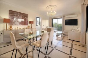 A modern living room with a glass dining table, white sofas, a chandelier, a flat-screen TV, and a large window. - FRGK Contemporary Villa StudioIndependent Pool (Le Cannet)